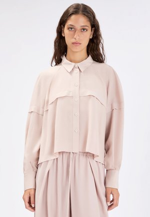 Giovane donna con capelli lunghi e ondulati che indossa una camicia larga color rosa pallido con bottoni e una gonna pieghettata abbinata su sfondo bianco.