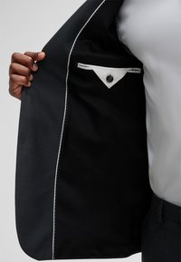 Das Innenfutter eines schwarzen Anzugsakts verfügt über eine glatte Innenverkleidung, eine weiße Tasche mit Knopf und eine Kontrastnaht entlang der Kante.