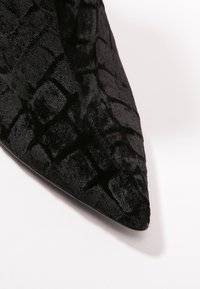 Zapato de terciopelo negro con punta afilada y una superficie texturizada que presenta un patrón geométrico. El material parece suave y aterciopelado.
