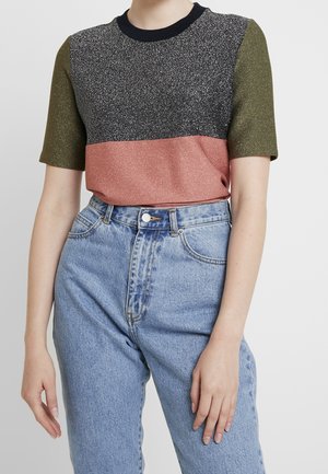 Mujer con una camiseta brillante de manga corta con bloques de color en oliva, negro y rosa, metida dentro de unos vaqueros de talle alto de color azul claro.