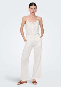 White, sleeveless jumpsuit with a button front, drawstring waist, and wide legs, made from a textured fabric. Worn with brown sandals.