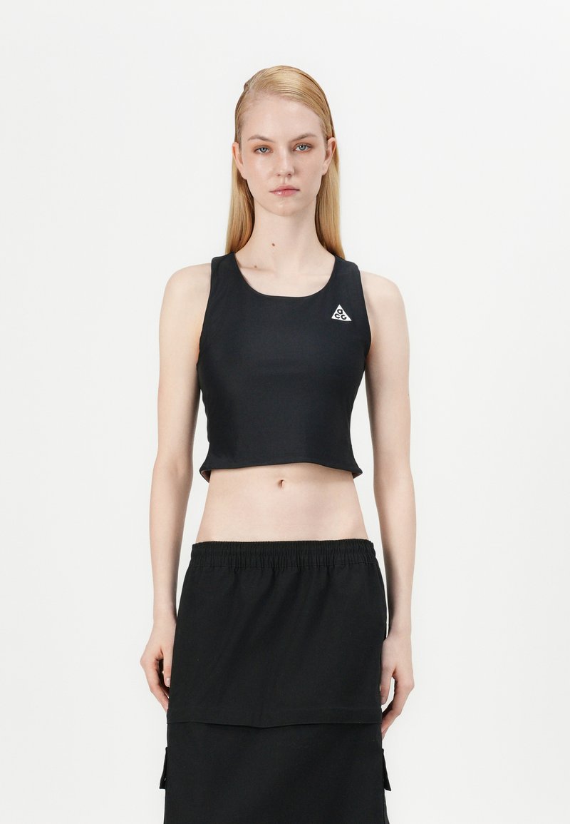 Melns īss tops no gludas auduma ar apaļu izgriezumu un nelielu trīsstūra formālo logotipu kreisajā pusē. Kopā ar melnu svārku.