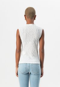 Hvit ermeløs topp med teksturert bølgedesign, kombinert med lyseblå tettsittende jeans. Bakvinkel fremhever plaggets form og design.