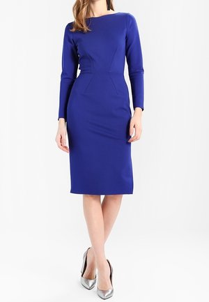 Femme portant une robe ajustée bleu roi longueur genoux à manches longues, associée à des escarpins argentés à bout pointu.