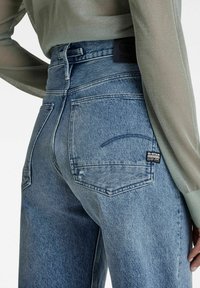 Jeans de mezclilla azul con cintura alta, lavado claro y bolsillos traseros. Presenta un parche de logo de cuero negro en la cinturilla.