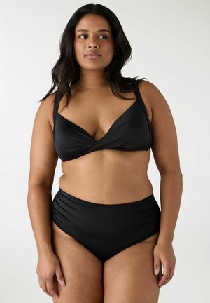 Donna che indossa un costume da bagno nero a due pezzi con un top stile incrociato e slip a vita alta, in piedi contro uno sfondo bianco semplice.