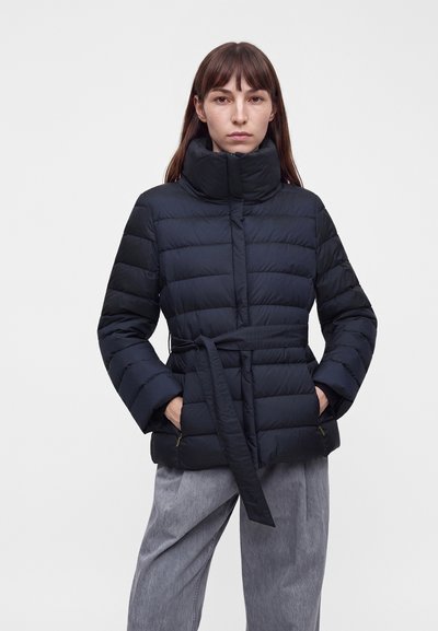 Femme portant une doudoune matelassée noire avec un col haut et une ceinture, associée à un pantalon gris ample, debout les mains dans les poches.