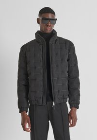 Crna puffer jakna s prešanim dizajnom, visokim ovratnikom i zatvaranjem na patentni zatvarač. Lagani materijal s glatkom teksturom i elastičnim manžetama.