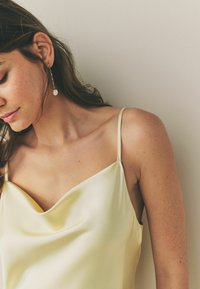 Satinová košile s cowl výstřihem v jemné žluté barvě s tenkými ramínky. Má hladkou texturu a elegantní splývavost, která zvýrazňuje linii krku.