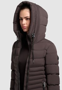 Padded brown jacket with a large, adjustable hood. Features a front zipper, ribbed accents, and a smooth exterior texture.