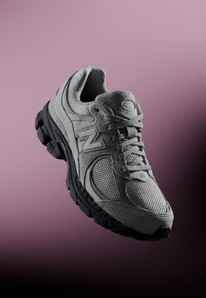 Chaussure de course New Balance grise avec tige en mesh et suède, semelle épaisse noire et lacets, sur un fond en dégradé du rose au noir.