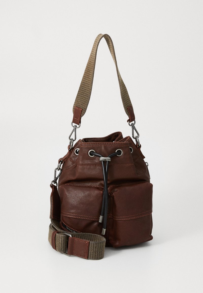 Brown leather bucket bag with front pockets, black drawstring closure, and wide beige fabric shoulder strap with leather accents.