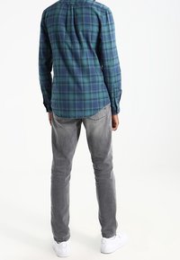 Man in een blauw en groen geruit shirt, grijze jeans en witte sneakers, staand met zijn rug naar de camera op een witte achtergrond.