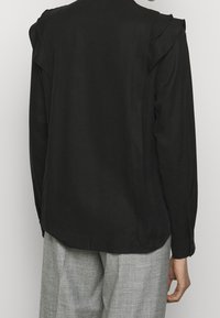Blouse noire avec détail de volants aux épaules, texture lisse, manches longues et coupe décontractée. Associée à un pantalon gris clair ajusté.