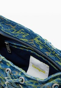 Torba dżinsowa z żółto-zielonymi wzorami, srebrnym zamkiem błyskawicznym, ściągaczem i białą etykietą wewnętrzną z nazwą marki „Desigual”.