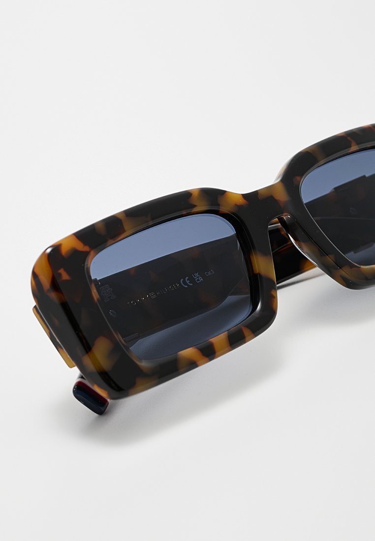 Lunettes de soleil en écaille de tortue avec des montures rectangulaires, des verres bleu foncé et une finition texturée. Présente un logo visible sur le bras intérieur.