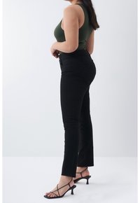 Femme debout de profil portant un haut vert sans manches, un pantalon noir ajusté avec des fentes aux chevilles et des escarpins noirs à brides et petit talon.