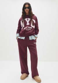 Felpa borgogna con grafica "NYC" e testo bianco, abbinata a pantaloni coordinati. Il modello indossa una camicia a righe sotto e scarpe slip-on beige.