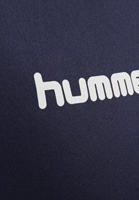 Zbliżenie białego napisu logo "hummel" na granatowym tle z tkaniny, pokazujące teksturowany materiał tkany.
