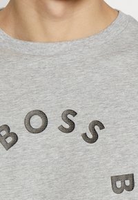 Detailní záběr na šedé tričko s kulatým výstřihem, na kterém jsou texturované černé písmena tvořící slovo "BOSS."