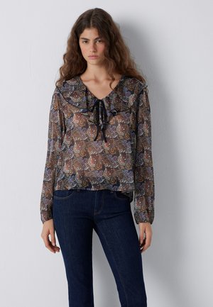 Frau mit langen, welligen Haaren, die eine transparente Paisley-Bluse mit Rüschenkragen und Bindeband trägt, kombiniert mit dunkelblauen Jeans, vor einer weißen Wand stehend.