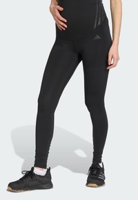 Leggings noirs avec une taille haute, fabriqu�és en tissu extensible. Présentent trois rayures diagonales sur le côté et sont associés à des chaussures de sport noires.