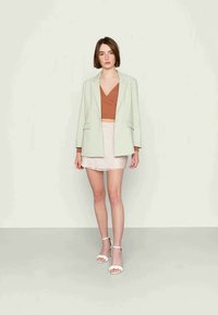 Blazer verde claro sobre uma blusa castanha e uma saia mini rosa com flores. Saltos altos completam o conjunto, apresentando uma textura suave em geral.