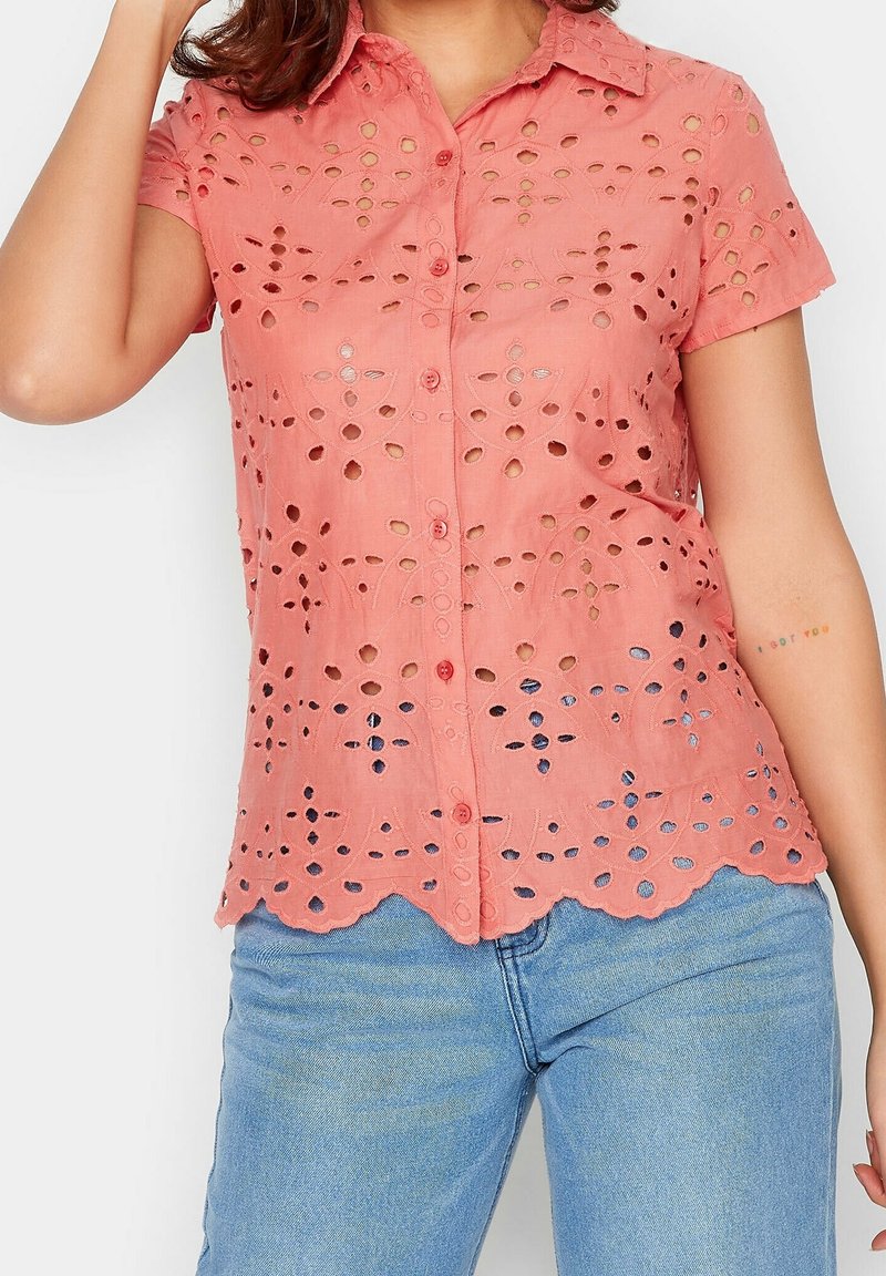Kobieta w koralowo-różowej bluzce z krótkim rękawem, z guzikami, i jasnoniebieskich dżinsach, z ręką przy głowie, widoczny tatuaż na przedramieniu.