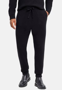 Czarne dzianinowe spodnie dresowe z regulowanym sznurkiem w pasie, zwężanymi mankietami i dwoma bocznymi kieszeniami. Noszone z czarnymi botkami za kostkę.
