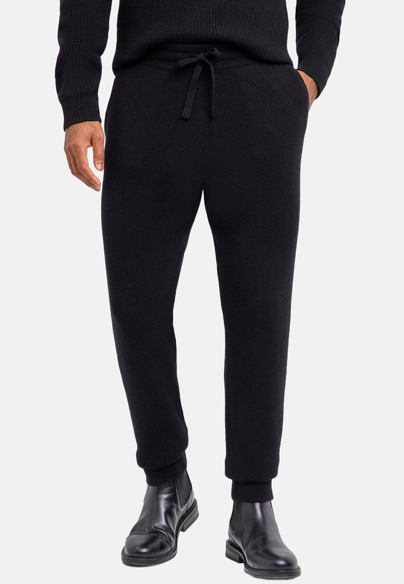 Czarne dzianinowe spodnie dresowe z regulowanym sznurkiem w pasie, zwężanymi mankietami i dwoma bocznymi kieszeniami. Noszone z czarnymi botkami za kostkę.