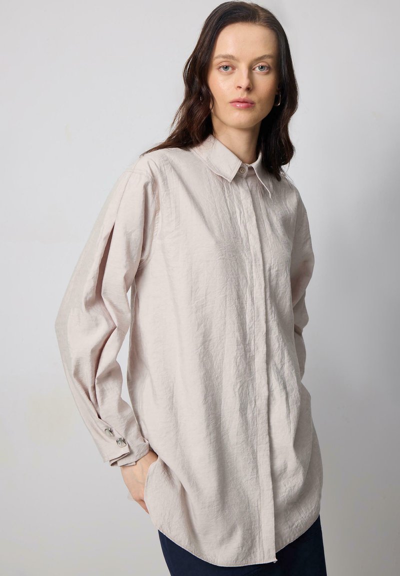 Light pink button-up shirt with a textured finish, long sleeves, and cuffs featuring round buttons. Loose fit with a rounded hem.