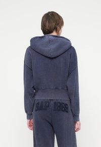 Navy skraćena hoodie s kapuljačom, s natpisom "GAP 1969" u teksturiranoj crnoj boji na leđima. Izrađena od mekanog pamuka.