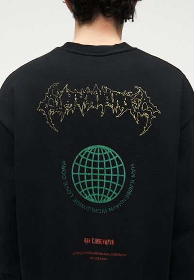 Άτομο που φοράει μαύρο φούτερ με στυλιζαρισμένο κίτρινο κείμενο, πράσινο γραφικό παγκόσμιου χάρτη και κόκκινο κείμενο που εμφανίζει λεπτομέρειες της μάρκας και του ιστότοπου στην πίσω πλευρά.