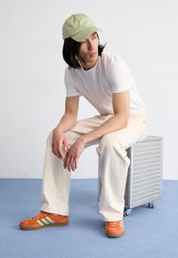 Green cap, white t-shirt, cream pants, and orange sneakers with teal stripes. Sitting on a silver metallic rolling cart against a plain backdrop.