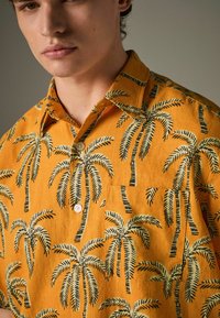 Kurzärmliges, kragenloses Hemd aus leuchtend orangefarbenem Stoff, mit einem Palmenmuster in Schwarz und Grün. Enthält eine Brusttasche und weiße Knöpfe.