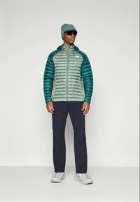 Man in een teal pufferjack, groene beanie, zwarte zonnebril, donkere broek en witte sneakers, staand tegen een effen achtergrond.