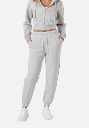 Kobieta w jasnoszarej, skróconej bluzie rozpinanej na zamek i dopasowanych dresach, w białych sneakersach na platformie, z rękami w kieszeniach, stoi na białym tle.