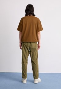 Tricou din bumbac maro, cu o croială lejeră, asortat cu pantaloni verzi măslinii. Converse albe vizibile pe picioare, stând pe un podea de culoare albastru deschis.