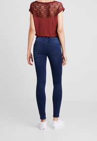 Jeans ajustados de color azul oscuro con una cintura de altura media, combinados con una blusa burdeos que presenta un respaldo de encaje transparente con un patrón de corazones. Zapatillas blancas.