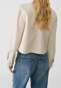 Frau mit langen blonden Haaren, die eine beige cropped Jacke und hoch taillierte blaue Jeans trägt, von hinten gezeigt.