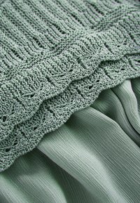 Světle zelená látka s pleteným topem se složitým krajkovým vzorem, vrstveným přes hladký, splývavý materiál s texturovaným povrchem.