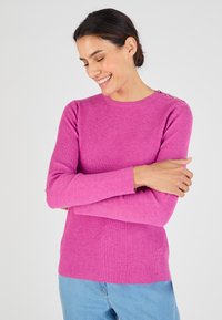 Aansluitende geribbelde trui in felroze met lange mouwen en knoopaccenten op de schouder, gecombineerd met lichtblauwe jeans.