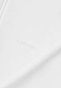 Tissu blanc présentant une texture douce, avec un logo brodé "LAMANIA" dans une couleur subtile et assortie. Design lisse et minimaliste.