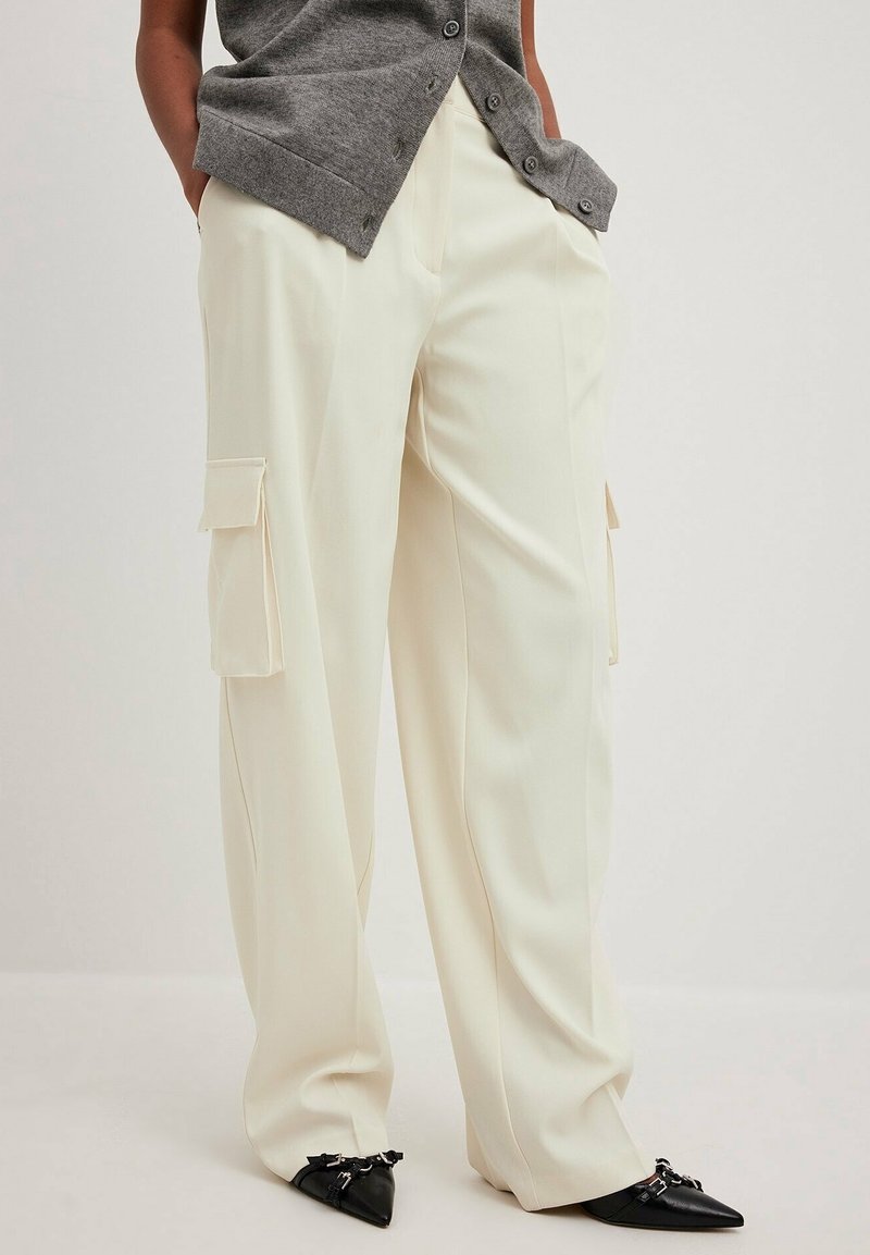 Personne portant un pantalon cargo crème à jambes larges avec poches latérales, un gilet gris boutonné, et des chaussures noires à bouts pointus avec boucles argentées.