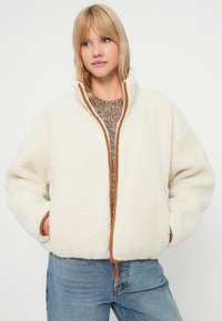 Krem boje flis jakna s visokim ovratnikom, smešnim detaljima na patentnom zatvaraču i bočnim džepovima. Nosi se preko pletene smeđe ljubavne.