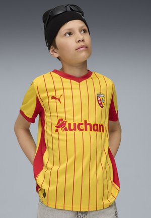 Un ragazzo indossa una maglietta da calcio a righe gialle e rosse con il logo Auchan, un berretto nero e occhiali da sole, mentre guarda in alto.
