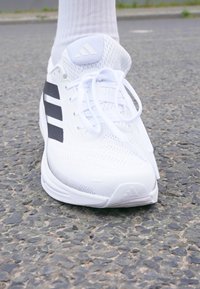 Baskets blanches avec tige en mesh, trois bandes noires sur le côté, bout arrondi et lacets blancs. Semelle texturée et logo sur la languette.