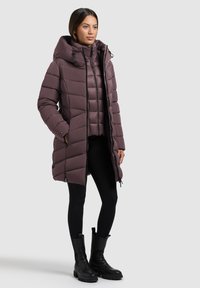 Dolga, prešita bunda v umirjeni mauve barvi s kapuco. Ima zadrgo, visok ovratnik in stranske žepe. Kombinirana s črnimi legicami in škornji.