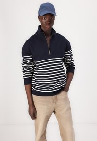 Navy quarter-zip pullover with white horizontal stripes, cotton blend fabric, long sleeves, and a kangaroo pocket. Paired with light beige pants and a blue cap.