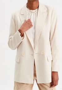 Beige blazer lavet af letvægtsstof, med enkeltknap-design, to forlommer og runde knapper i en matchende farve.